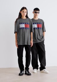 Camisetas de gran tamaño en gris con un logo de Tommy Jeans en azul marino, rojo y blanco en la parte frontal, combinadas con pantalones negros y zapatillas.