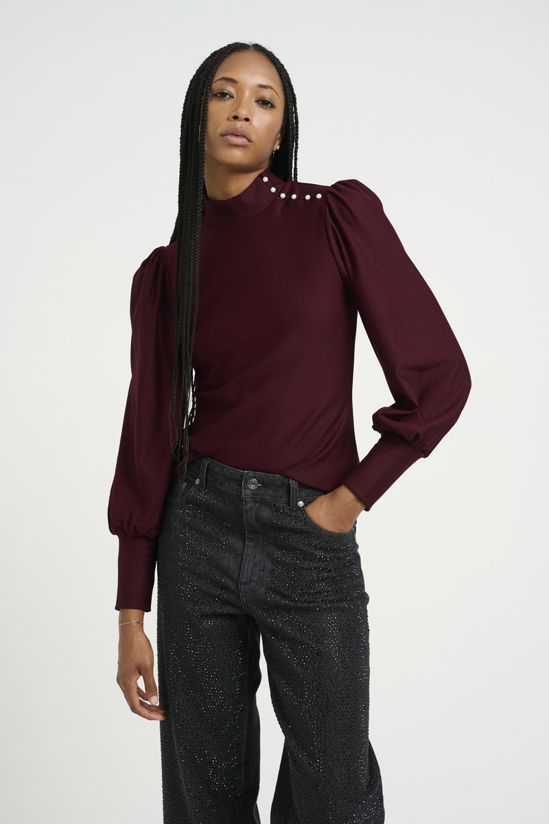 Bordeaux langærmet top med høj hals og puffede ærmer, accentueret med hvide dekorative knapper på skuldrene. Båret med glitrende mørke jeans.