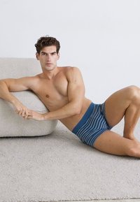 Blaue gestreifte Boxershorts aus weichem Material mit einem einfarbigen Bund, präsentiert von einem Mann, der sich auf einer strukturierten beigen Oberfläche entspannt.