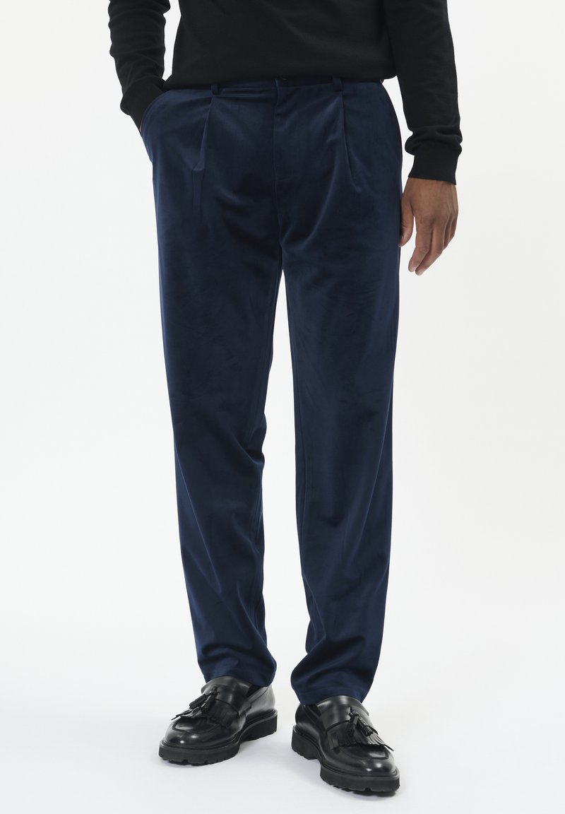 Tamno plave baršunaste hlače s opuštenim krojem, dva prednja nabora i funkcionalnim bočnim džepovima. Nosene s crnim cipelama.