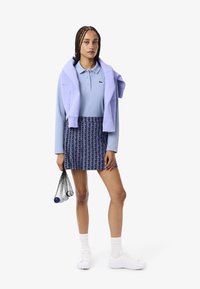 Polo shirt lilla, maglione lilla lavorato a maglia drapes sulle spalle, gonna navy a fantasia, sneakers bianche e borsa nera in rete con palline.