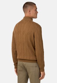 Brauner Strickpullover mit einem erhabenen Muster. Verfügt über einen hohen Kragen und gerippte Bündchen. Der Stoff wirkt weich und strukturiert.