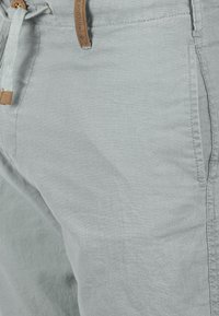 Pantalones de lino gris claro con un acabado texturizado, que cuentan con una cintura con cordón y un acento de cuero. Los bolsillos son visibles a los lados.