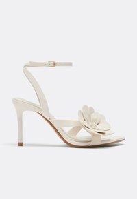 Sandale à talons hauts beige avec bride à la cheville et grande décoration florale sur la bride des orteils, présentée sur un fond neutre.