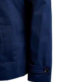 Zunanja oblačila mornarsko modre barve iz gladkega, vodoodpornega blaga. Vključuje žepek za vstavljanje in detajl z gumbom na manšeti za dodatno funkcionalnost.