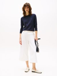 Kvinne iført en marineblå langermet topp, hvite vide bukser med cropped lengde, hvitt belte, loafers og holder en liten svart håndveske.