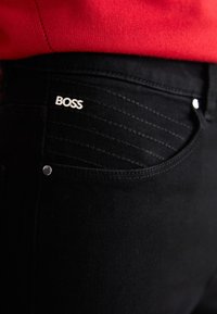 Jeans negros con detalles de bolsillo visibles, rayas texturizadas en la cadera y un logo plateado "BOSS" en el bolsillo delantero. Acompañados de una blusa roja.