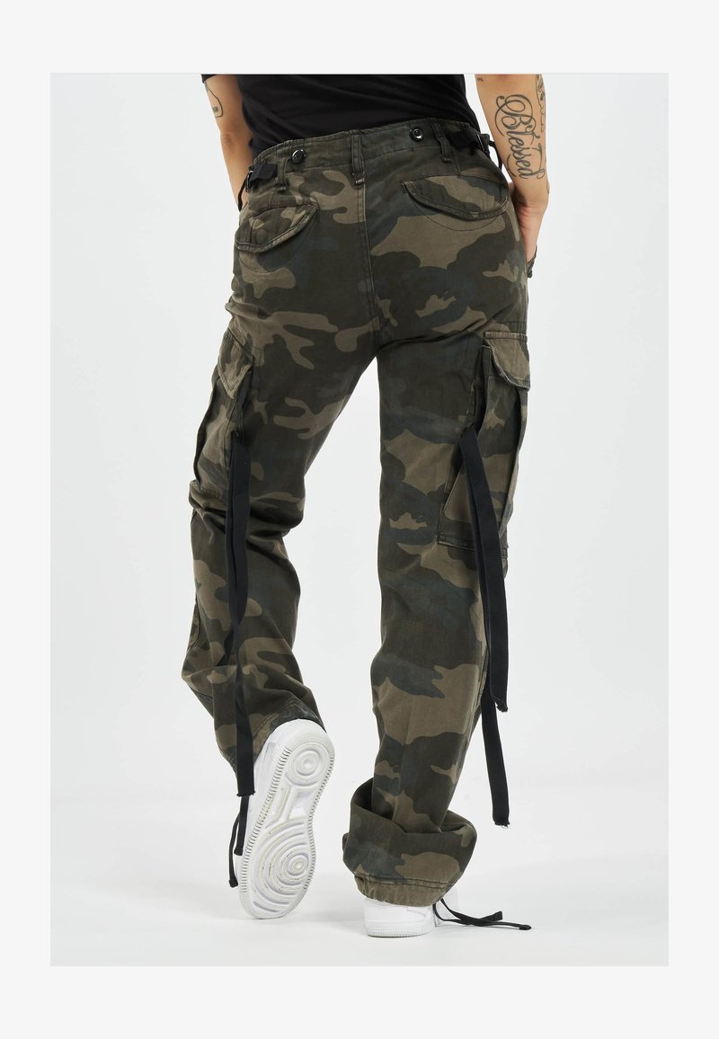 Osoba koja nosi široke cargo hlače u maskirnom uzorku s crnim trakama koje vise, uparene s bijelim tenisicama, vidi se odostraga.