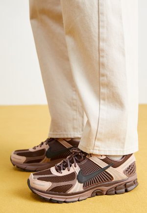 Nike-Sneaker sind aus einer Mischung aus braunem und hellrosa Netz- und Gummi-Material gefertigt, mit schwarzen Akzenten und einer strukturierten Sohle, kombiniert mit beigen Hosen.