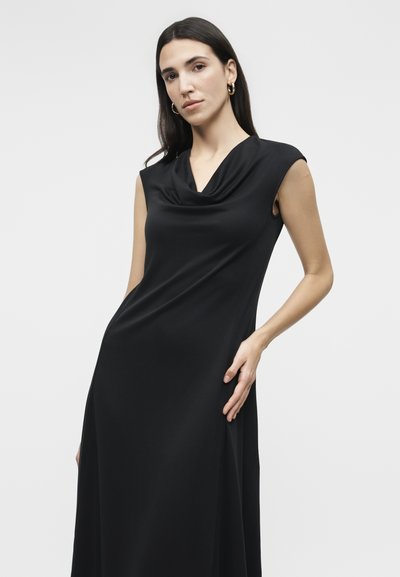 Γυναίκα με μακριά σκουρόχρωμα μαλλιά που φοράει χρυσούς κρίκους στα αυτιά και μαύρο φόρεμα χωρίς μανίκια με ντραπέ λαιμόκοψη, ποζάροντας με το ένα χέρι στη μέση της.