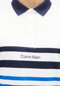 Λευκό πόλο μπλουζάκι με γιακά σε ναυτικό μπλε και οριζόντιες ρίγες σε ναυτικό και μπλε, με το λογότυπο "Calvin Klein" στο στήθος.