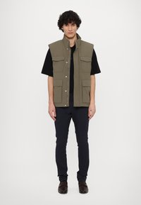 Olivengrøn vest med høj krave, flere frontlommer og trykknapper, lagt over en sort skjorte, parret med marineblå bukser.