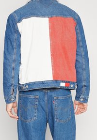 Person som bär blå denimjeans och en blå denimjacka med vita och röda färgblock samt en Tommy Jeans-logotyp på ryggen.