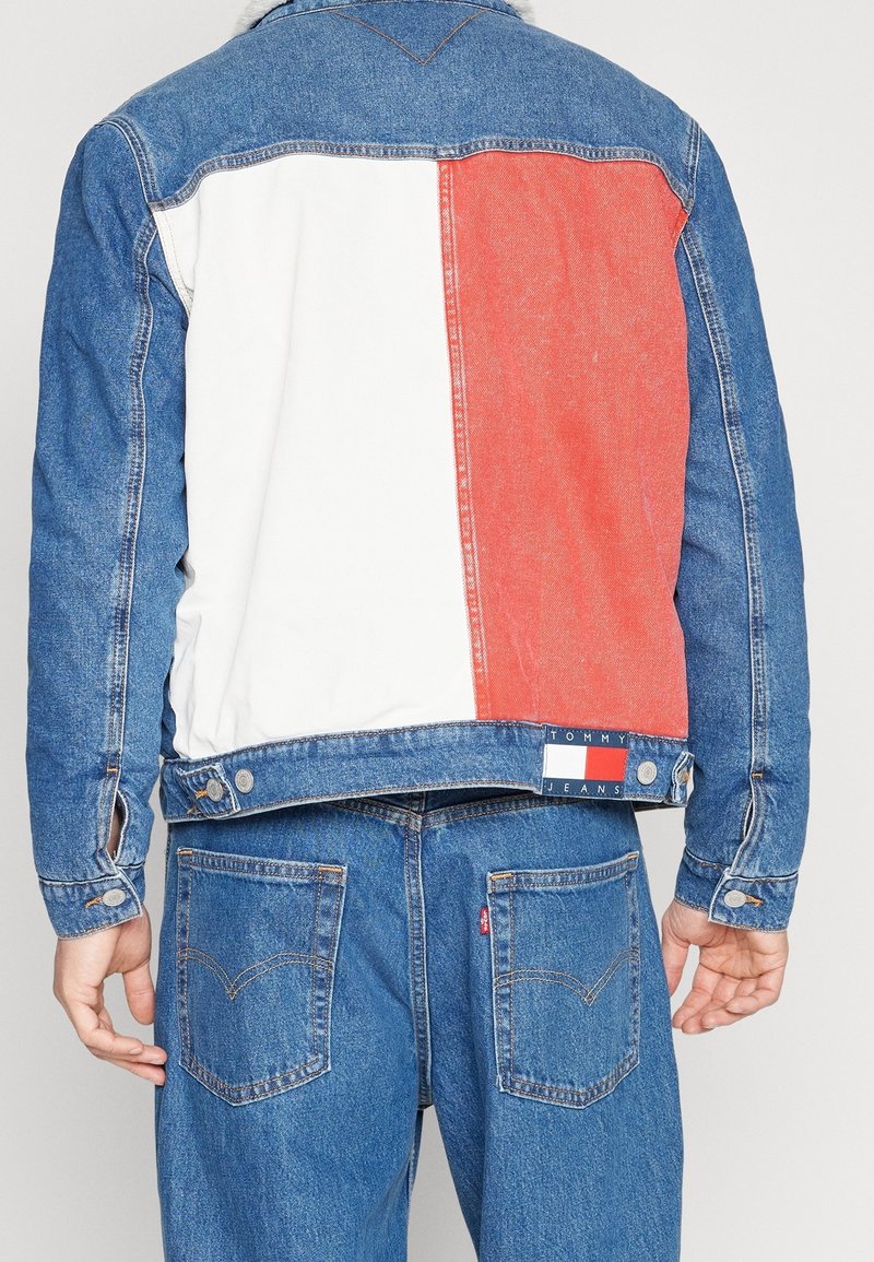 Person som bär blå denimjeans och en blå denimjacka med vita och röda färgblock samt en Tommy Jeans-logotyp på ryggen.