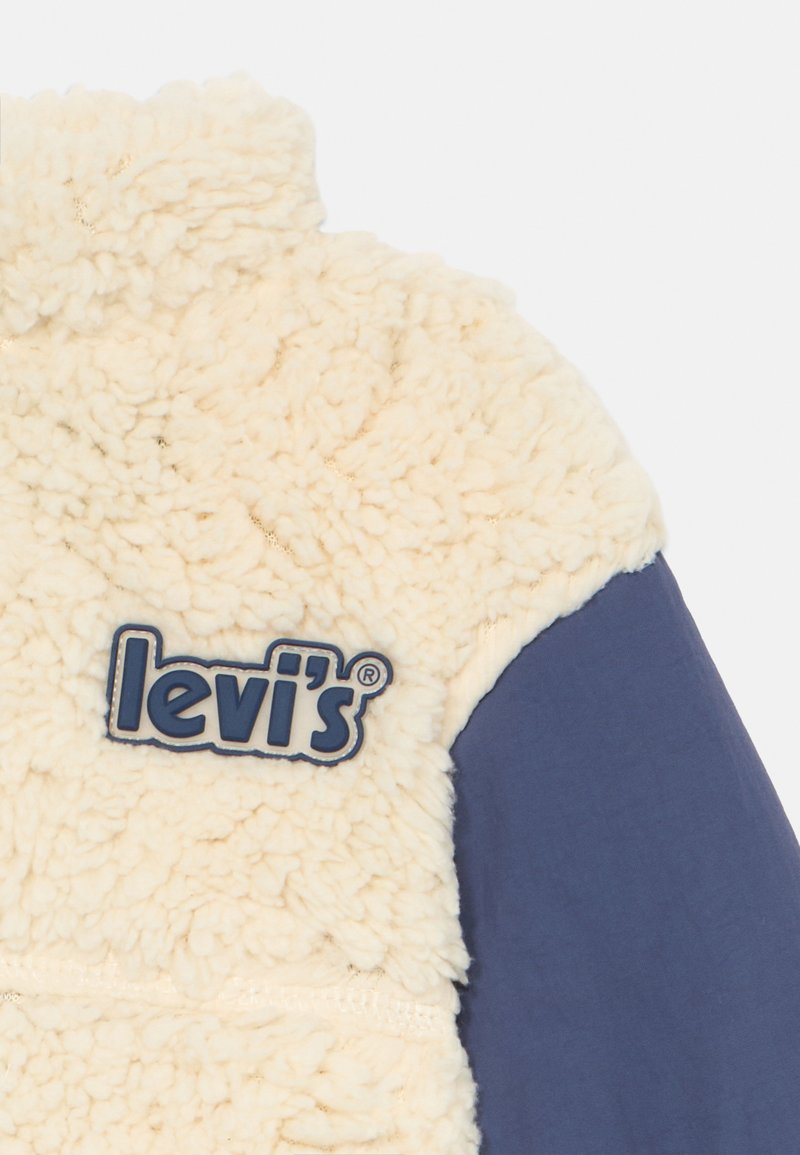 Chaqueta de forro polar tipo sherpa crema con mangas azul marino. Presenta un destacado logo azul de Levi's en el pecho y un tejido texturizado.
