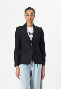 Černý blazer se dvěma knoflíky, s klopami a předními kapsami, oblečený přes bílou košili. Doplňuje ho světle modré džíny.