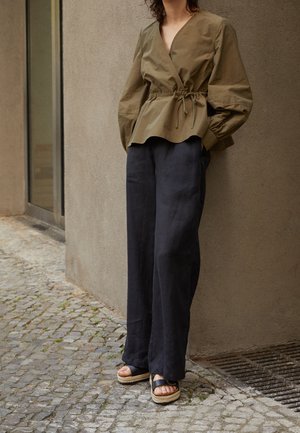 Personne portant un top cache-cœur olive avec une taille à cordon, un pantalon noir à jambes larges et des sandales plateformes noires, debout sur un pavé.