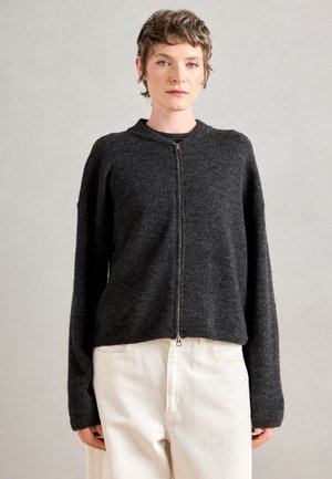 Pull zippé gris foncé avec un col rond, texture douce, coupe ample et manches longues, associé à un pantalon blanc.