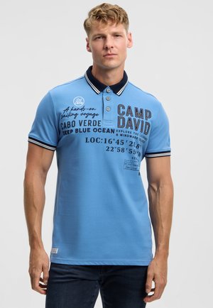 Hellblaues Poloshirt mit marineblauem Kragen und gestreiften Kanten, mit gedrucktem Text und Designs. Hergestellt aus weichem Stoff, kurze Ärmel, Knopfleiste.