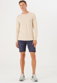 Langärmliges, hellbeiges Hemd mit strukturiertem Design, kombiniert mit dunkelblauen, hochgerollten Shorts. Das Schuhwerk hat helle blaue Akzente und Schnürsenkel.