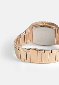 Roségoldene Metalluhr mit rechteckigem Gehäuse, verbundenem Armband und Schnallenverschluss. Rückseite zeigt Markeninformationen und Spezifikationen.