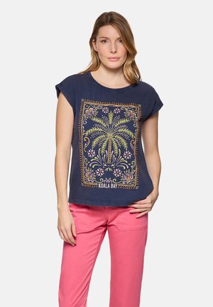 Mujer vistiendo una blusa sin mangas azul marino con un estampado colorido de palmeras y pantalones rosas, de pie frente a un fondo liso.