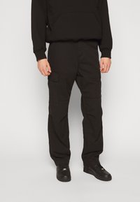 Schwarze Cargo-Hosen aus strapazierfähigem Stoff, mit Seitentaschen, lockerer Passform und flachem Frontdesign, kombiniert mit schwarzen Sneakers.