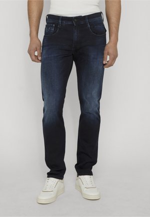 Uomo che indossa jeans slim-fit scuri lavati e sneakers bianche, in piedi davanti a uno sfondo chiaro e uniforme.