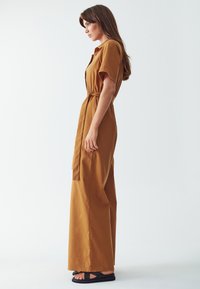 Hnědý bavlněný overal s krátkými rukávy, knoflíkovým zapínáním vpředu, opaskem kolem pasu a širokými nohavicemi, který je nošen s černými sandály.