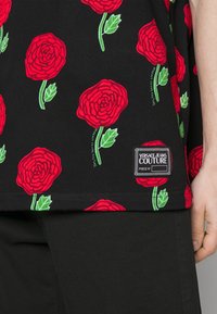 Black shirt with red rose prints and green leaves, featuring a Versace Jeans Couture label near the hem, worn with black pants.