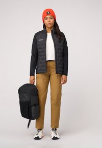 Svart vadderad jacka med åtsittande ärmar, orange stickad mössa, tan färgade byxor och en svart ryggsäck. Vita sneakers med svarta detaljer bärs.