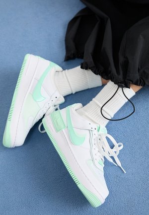 Baskets blanches et vert menthe portées avec des chaussettes blanches côtelées et un pantalon noir avec des cordons réglables sur un sol moquetté bleu.