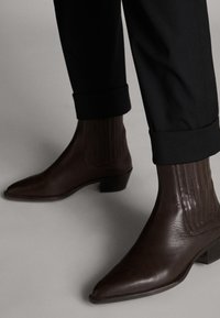 Braune Leder-Stiefeletten mit spitzen Zehen und strukturierten Seitenpaneelen. Niedrige Blockabsätze; getragen zu schwarzen, umgeschlagenen Hosen.