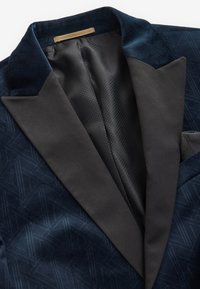 Blazer en velours bleu marine avec revers en satin noir à pointe et doublure intérieure à motif nid d'abeille, montrant le col et la poitrine.