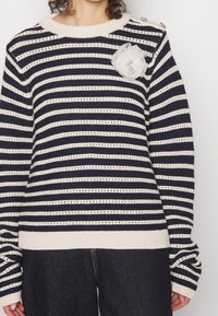 Pull en tricot rayé bleu marine et crème, avec un col rond, des manches longues, un accent en dentelle en forme de fleur et des boutons décoratifs aux épaules.