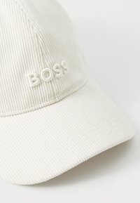 Witte corduroy pet met een gebogen rand, geborduurd "BOSS" logo in lichte kleuren en een ribbelstructuur door de stof.