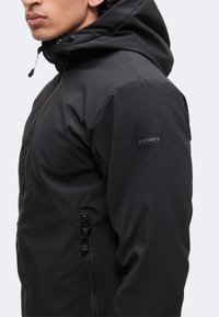 Vue de côté d'une personne portant une veste noire à capuche avec une poche zippée sur le bras et un patch logo sur la manche.