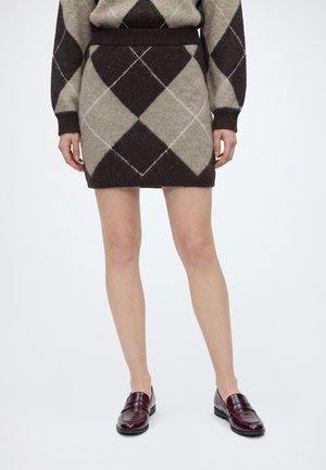 Femme portant une jupe en tricot argyle marron et beige et un pull assorti avec des mocassins bordeaux, debout devant un fond blanc uni.