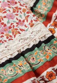 Kolorowa tkanina z wzorami w kwiaty w kolorach czerwonym, różowym i zielonym; ma koronkowe wykończenie oraz warstwowe, falbaniaste detale.