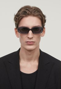 Gafas de sol con un elegante marco de plástico morado oscuro, lentes rectangulares y brazos finos, llevadas por un hombre con un blazer y una camisa negros.