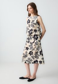 Bloemmotief midi-jurk in lichtbeige en donkerblauw, met een losse pasvorm, mouwloos ontwerp en een elastische taille. Zwarte instap schoenen.