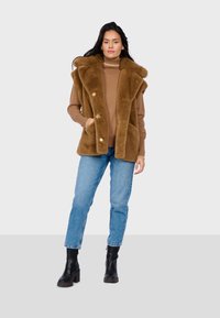 Gilet in finta pelliccia marrone con cappuccio, bottoni frontali e tasche laterali, abbinato a un dolcevita beige, jeans blu e stivaletti neri.