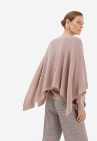 Světle růžový volně padnoucí pončo svetr s ribbovanou strukturou, vrstveným designem a širokými rukávy, doplněný kostkovanými, dobře padnoucími kalhotami.