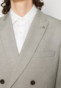Světle šedý dvouřadý blejzr s herringbone vzorem. Obsahuje náprsní kapsu a tmavé knoflíkové detaily. Nositelný přes bílou košili.