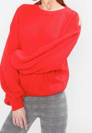 Femme portant un pull en tricot rouge vif avec une épaule découpée et un pantalon ajusté à carreaux gris, posant avec une main détendue.