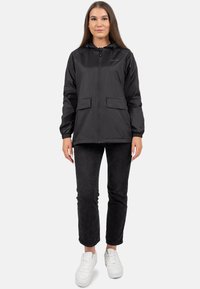 Giacca leggera nera con zip frontale, cappuccio regolabile e due tasche anteriori, abbinata a jeans scuri e sneakers bianche.