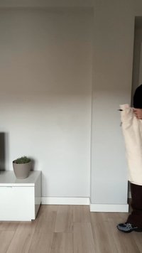 Asmuo, laikantis kremine spalva išvyniotą kilimą, stovintis ant šviesios medinės grindų šalia balto kabineto su vazonine augalu.