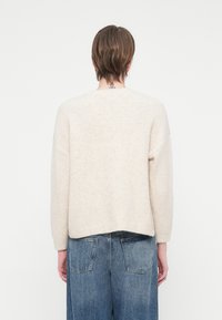 Cilvēks ar īsiem brūniem matiem, tērpies bēšā adītā džemperī un zilos džinsos, stāv atgriezies pret baltu fona.