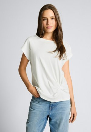 Młoda kobieta z długimi brązowymi włosami, ubrana w biały top z krótkim rękawem i niebieskie dżinsy, stojąca z jedną ręką w kieszeni na jednolitym tle.