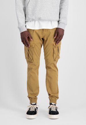 Tan cargobroek met elastische boorden, uitgerust met meerdere cargopockets, gecombineerd met zwarte sneakers en een grijze sweatshirt over een witte top.